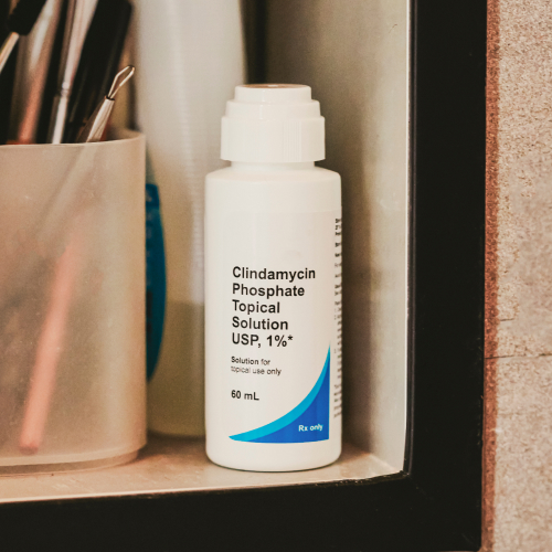 A pristine white bottle of Clindamycin Phosphate Topical Solution, 1% USP, is neatly placed on a shelf alongside a container brimming with pens and other essentials.