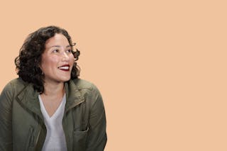 Woman with curly hair in a green jacket and white shirt smiling while looking to the left, against a plain beige background.