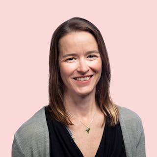 A smiling woman with long brown hair, wearing a black top and gray cardigan, on a plain light pink background.