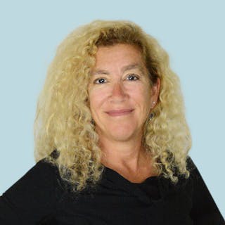 A person with curly blonde hair smiles, wearing a black top, standing against a light blue background.