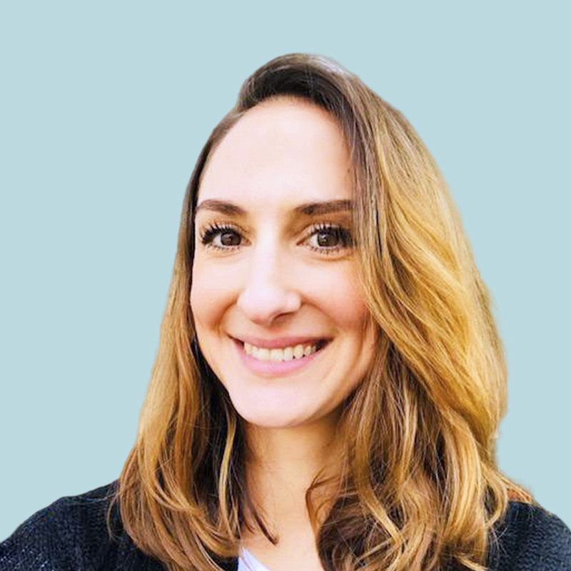 A person with shoulder-length brown hair smiles in front of a plain greenish background.