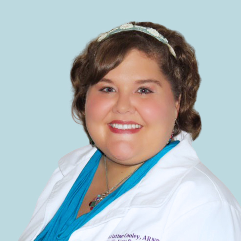 Person with short brown hair, wearing a white coat and blue top, smiling against a light blue background.