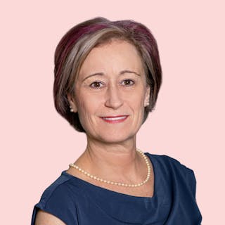 A person with short hair, wearing a blue top and a pearl necklace, smiling against a pink background.