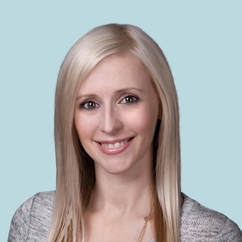 A person with long blonde hair in a gray top smiles at the camera against a light blue background.