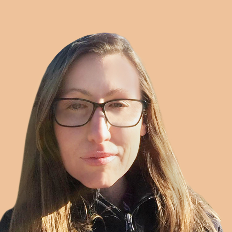 A person with long brown hair and glasses is wearing a black jacket against a plain peach background.