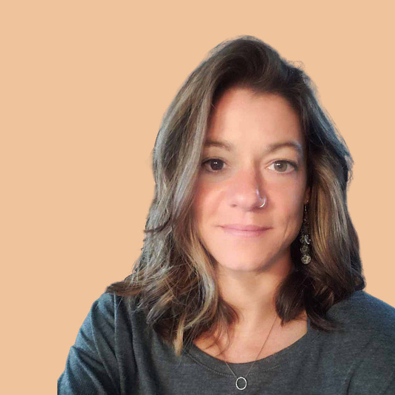 Person with shoulder-length hair and a nose ring, wearing a gray shirt, against a solid beige background.