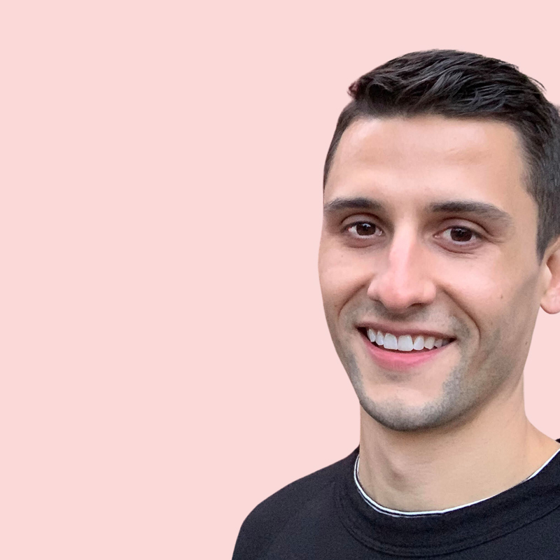 A person with short dark hair and a black shirt smiles against a pink background.