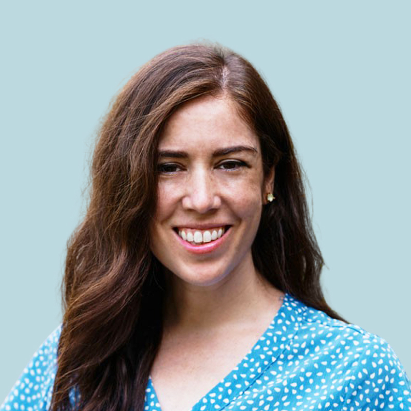 A person with long brown hair smiles, wearing a blue polka dot shirt against a plain light blue background.