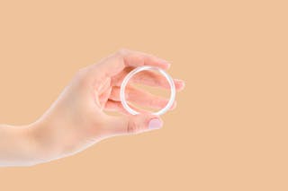 A hand holding a clear, round contraceptive ring against a beige background.