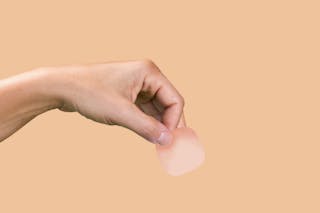 A hand holding a small, square, translucent adhesive patch against a solid peach background.