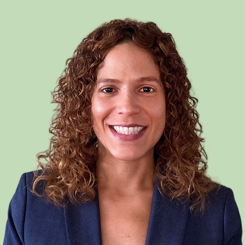 Woman with curly hair wearing a navy blazer smiles against a light green background.