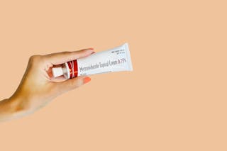 Hand holding a tube of Metronidazole Topical Cream 0.75% against a peach-colored background.