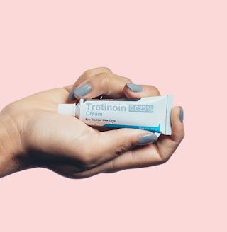A hand holds a tube of Tretinoin 0.025% cream against a pink background.