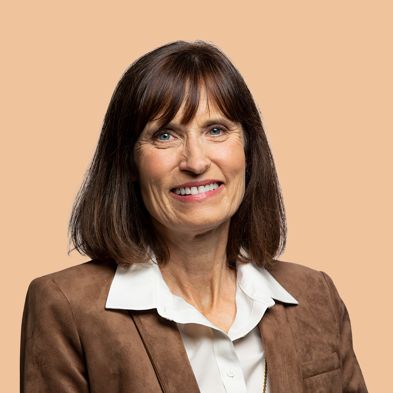 A woman with shoulder-length brown hair smiles, wearing a brown blazer over a white shirt against a tan background.