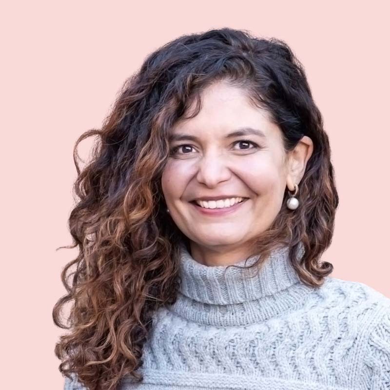 Mujer con cabello rizado sonriendo, vistiendo un suéter gris sobre un fondo rosa.