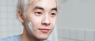 A person with light hair applies skincare cream to their face, standing in a tiled bathroom.