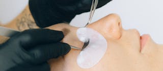 Close-up of an eyelash extension application, showing gloved hands using tweezers to attach lashes to a person's eyelid.