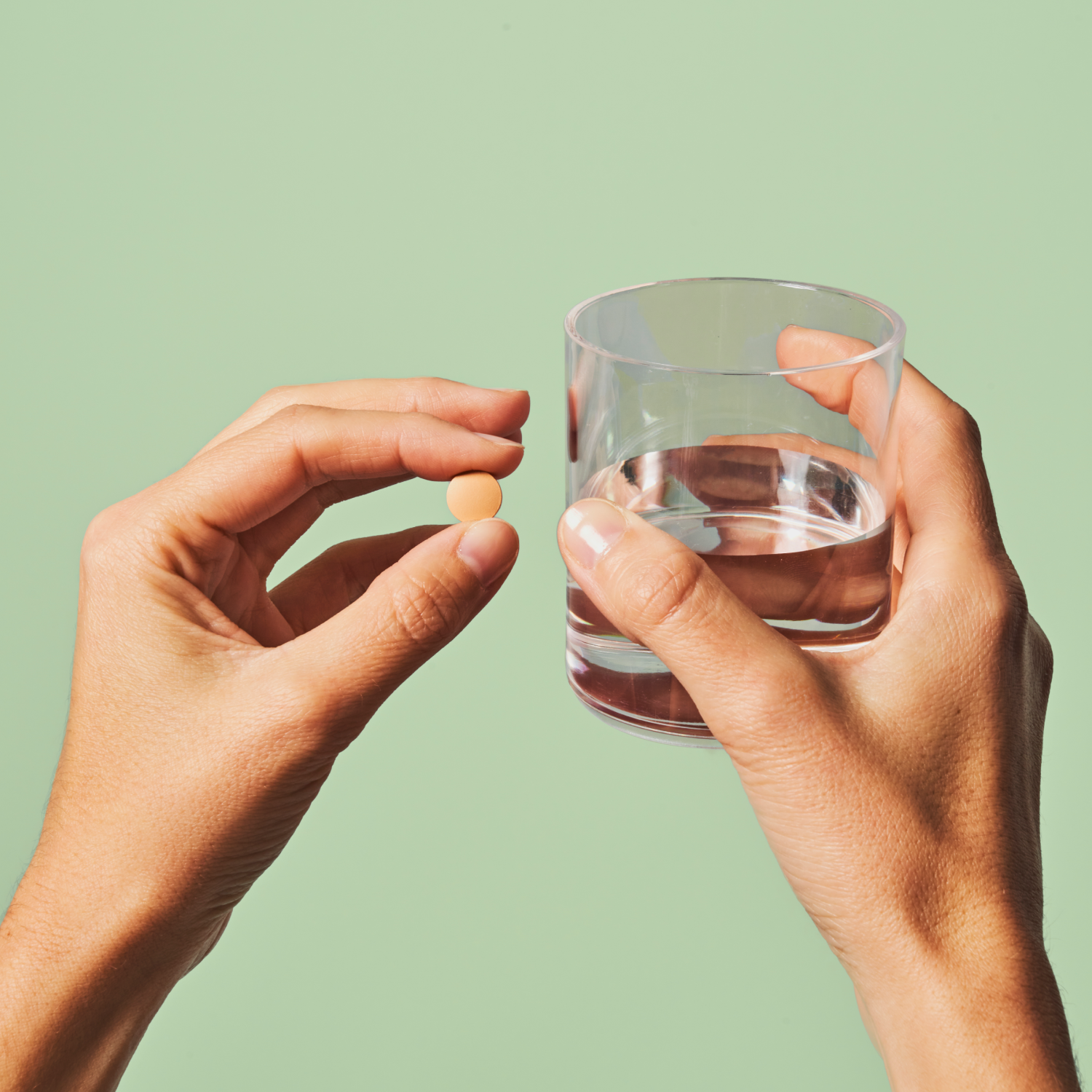 A person holding a glass of water in their right hand and a small round pill in their left hand against a green background.