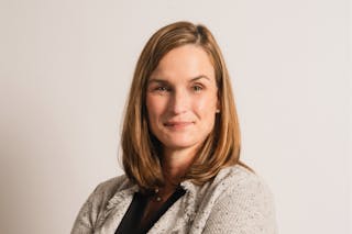 A woman with shoulder-length brown hair is wearing a light gray blazer over a black top, standing against a plain white background.