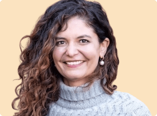 A person with long curly hair, wearing a gray turtleneck sweater, smiles against a plain background.