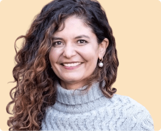 A person with long curly hair wearing a gray turtleneck sweater smiles against a beige background.