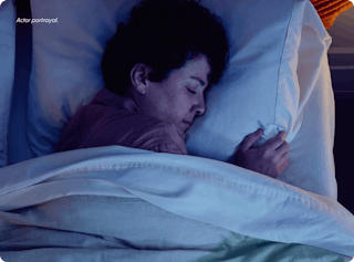 Person sleeping peacefully in bed with white pillows and blanket, illuminated by soft light.
