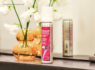 A bottle of Minoxidil topical aerosol foam for women on a bathroom counter, next to an orange vase with white flowers.