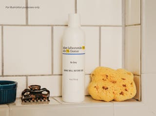 A bottle of sodium sulfacetamide and sulfur cleanser is placed on a tiled bathroom shelf, alongside a yellow sponge, a black hair clip, and a small blue dish.
