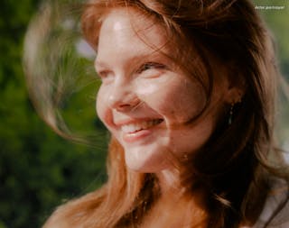 Woman with red hair smiling outdoors with blurred green background.