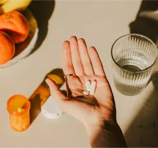 A hand holds two white pills. Nearby are an open prescription bottle and a glass of water. A plate of oranges and bananas is in the background.