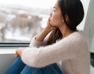 Person sitting by a window, gazing outside with head resting on hand, wearing a soft sweater and jeans.