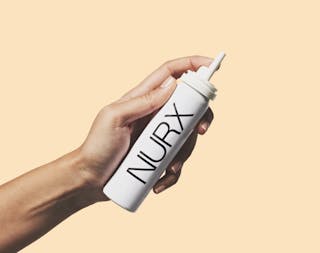 A hand holding a white aerosol can labeled “NURX” against a beige background.