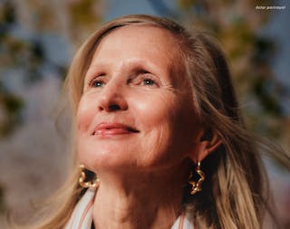 A woman with long blond hair smiles while looking upwards. She is wearing star-shaped earrings and a striped scarf, with a blurred background of trees.