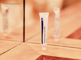 Un tubo de crema de tretinoína, USP 0.1% se encuentra sobre una superficie con textura de madera, con un reflejo de espejo en el fondo.