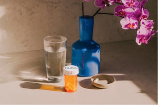 A prescription pill bottle, a glass of water, a blue vase with purple flowers, and a small dish are placed on a table.