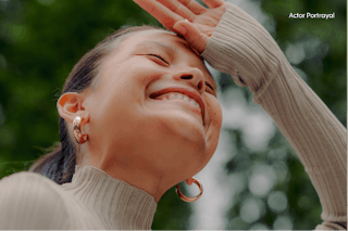 A person in a beige sweater and hoop earrings smiles with eyes closed against a blurred green background.