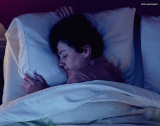 Person with short curly hair sleeping peacefully in bed, holding a pillow, under white sheets.