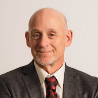 Bald man with a warm smile wearing a gray suit jacket, white shirt, and red plaid tie against a plain background.