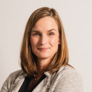 A person with shoulder-length brown hair and wearing a light gray blazer smiles at the camera against a plain background.