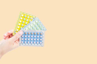 A hand holds three packs of birth control pills against a plain background.