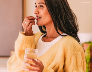 A woman in a yellow sweater stands in a well-lit room, holding a glass of water as she takes her UTI medication.