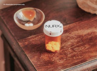 Prescription pill bottle with white and yellow tablets on a wooden surface. Bowl with two white pills and an orange wedge in the background.