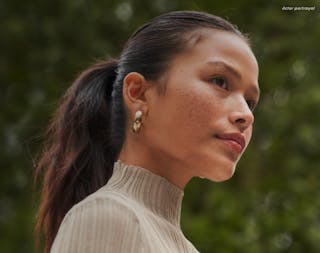 A person with long dark hair in a ponytail, wearing a ribbed beige top and gold hoop earrings, looks to the side.