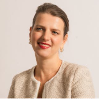 Smiling person with a light blazer and red lipstick against a plain background.