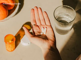 Hand holding two white pills, with a pill bottle, blister pack, and a glass of water on a table nearby.