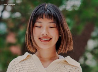 Person with short hair and a white top smiles while looking down, with greenery in the background.