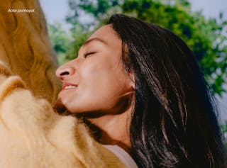 A person in a yellow sweater smiles with eyes closed, facing upward against a backdrop of blurred greenery. Text reads, 