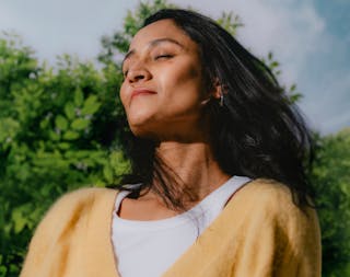 A person with long dark hair and a yellow sweater stands outdoors, eyes closed, facing the sunlight, with greenery in the background.