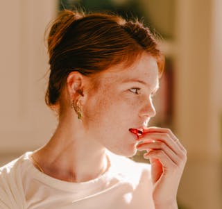 A person with red hair and freckles looks thoughtfully to the side, wearing a white shirt and earrings, with sunlight on their face.