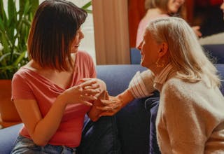 Two women sit on a couch engaged in conversation, with one smiling. A large plant is in the background.